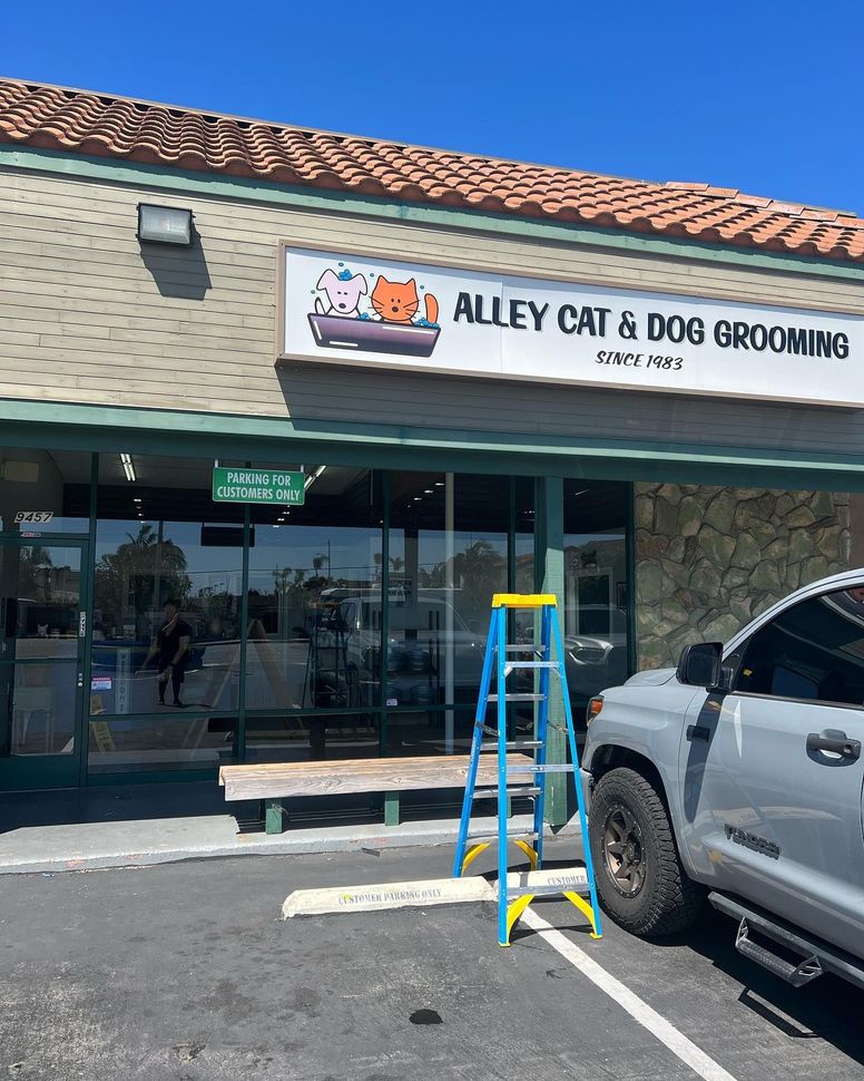 Storefront sign for Alley Cat & Dog Grooming with a ladder in front and a parked truck nearby.