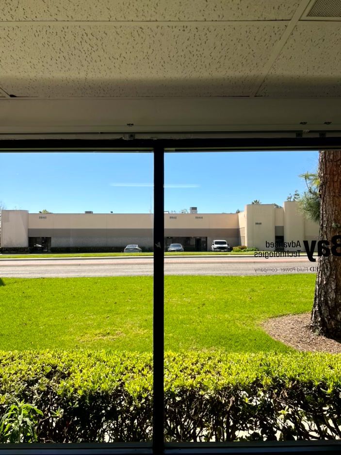 View through a window of a green lawn, hedge, road, and beige building on a sunny day