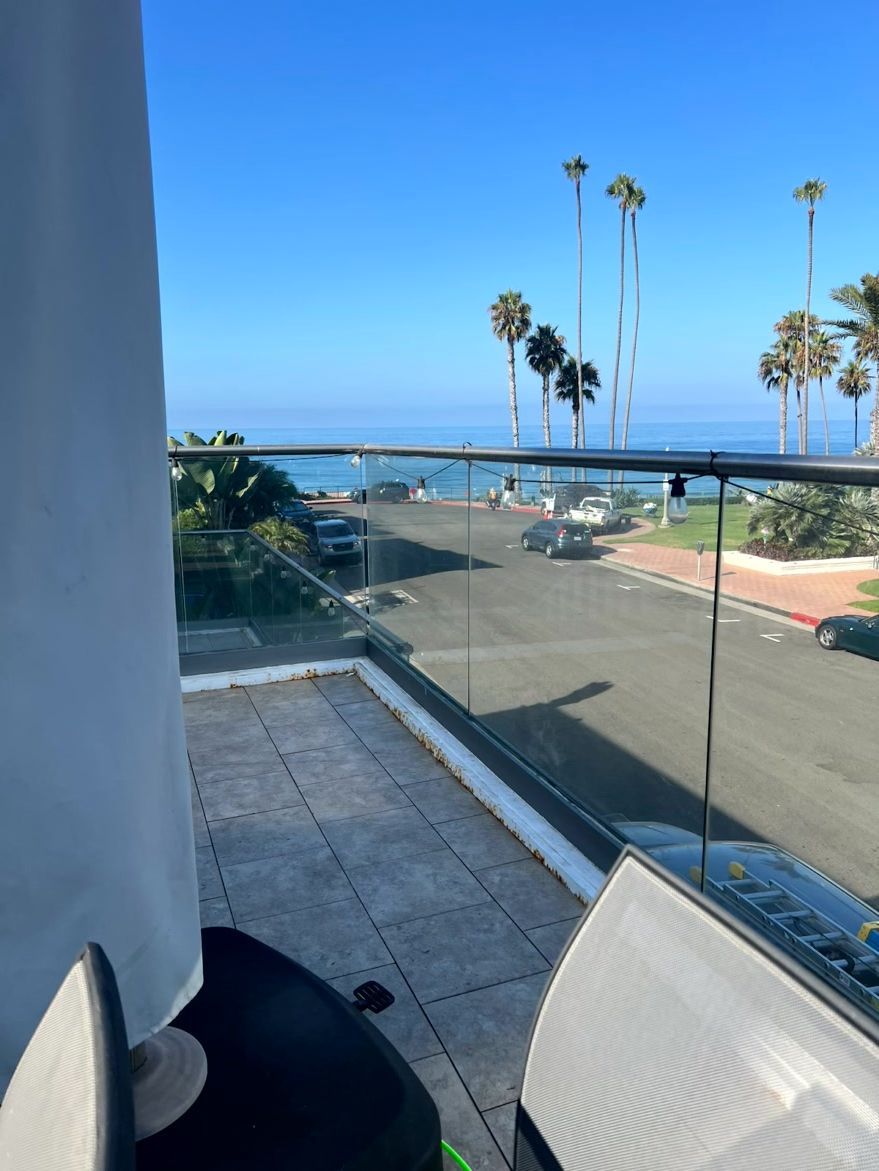 Balcony with glass railing overlooking a road, palm trees, and blue sky near the coast