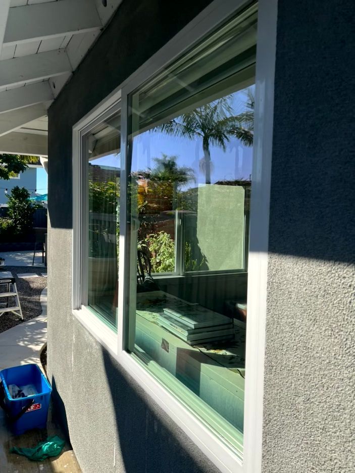 Exterior view of a house window with white trim, reflecting palm trees and a sunny yard.