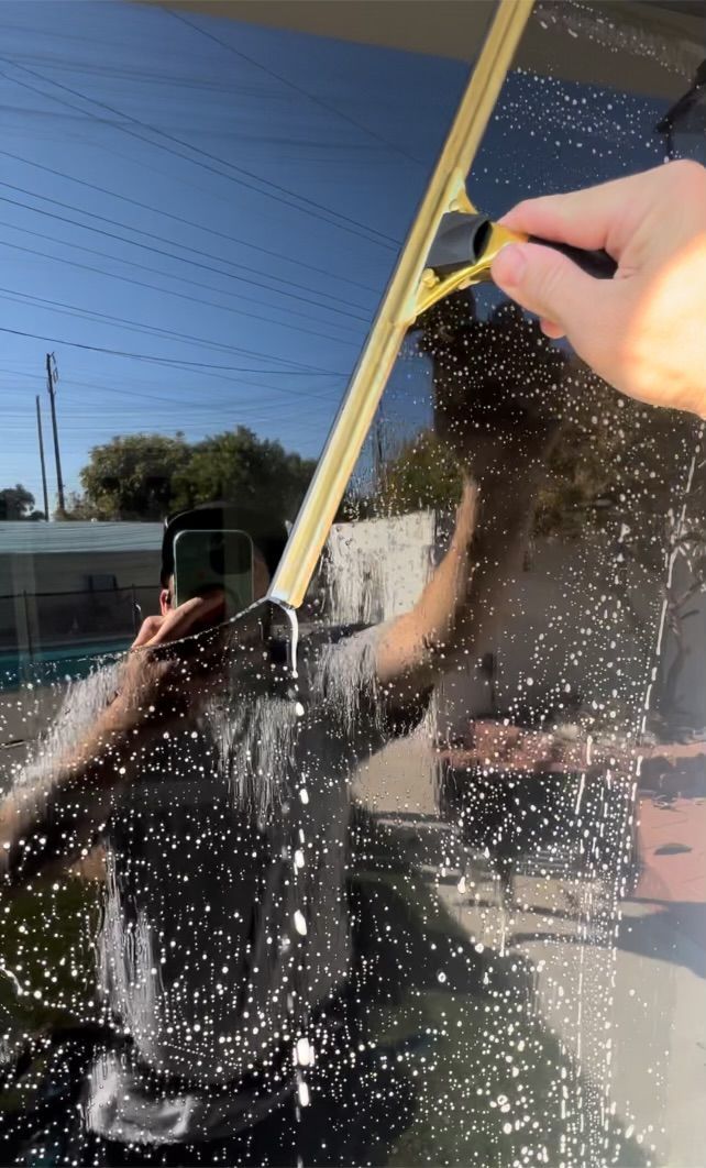 Person cleaning a glass window with a squeegee, water droplets splashing in sunlight.