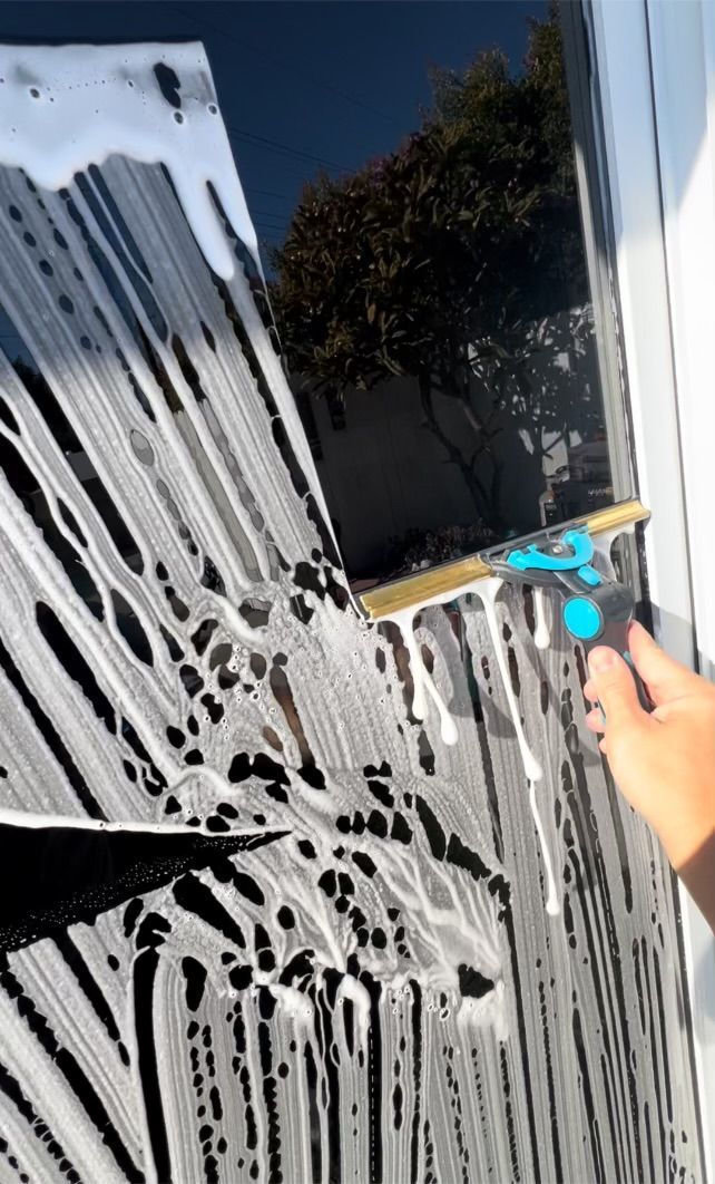 Hand using a blue squeegee to clean soap-covered glass window outdoors