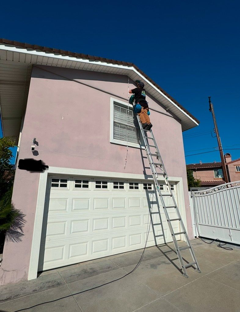 Worker on a ladder painting the upper wall of a pink garage under a clear blue sky