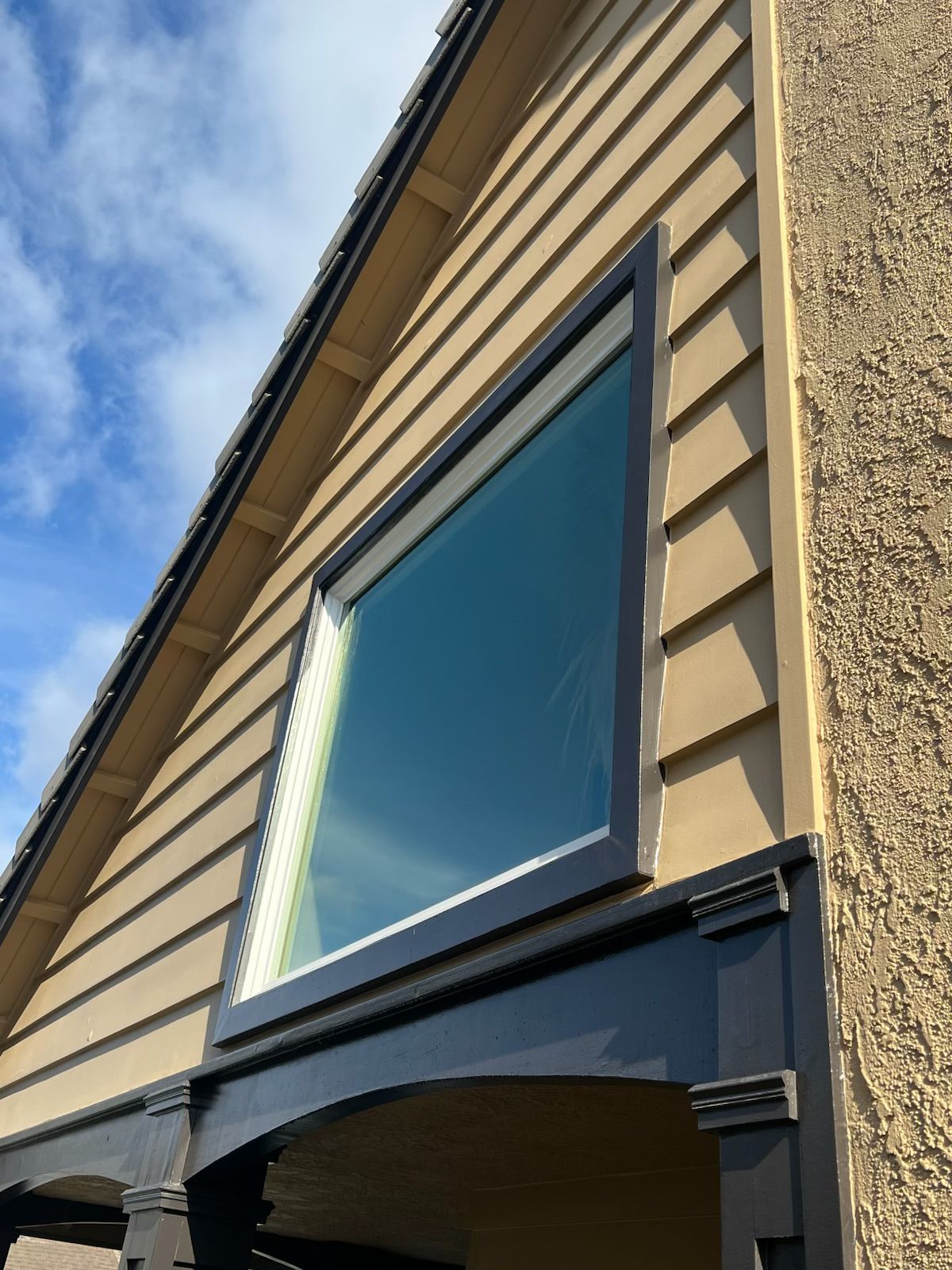 House exterior with tan siding, a large blue-tinted window, and part of a blue awning against a cloudy sky