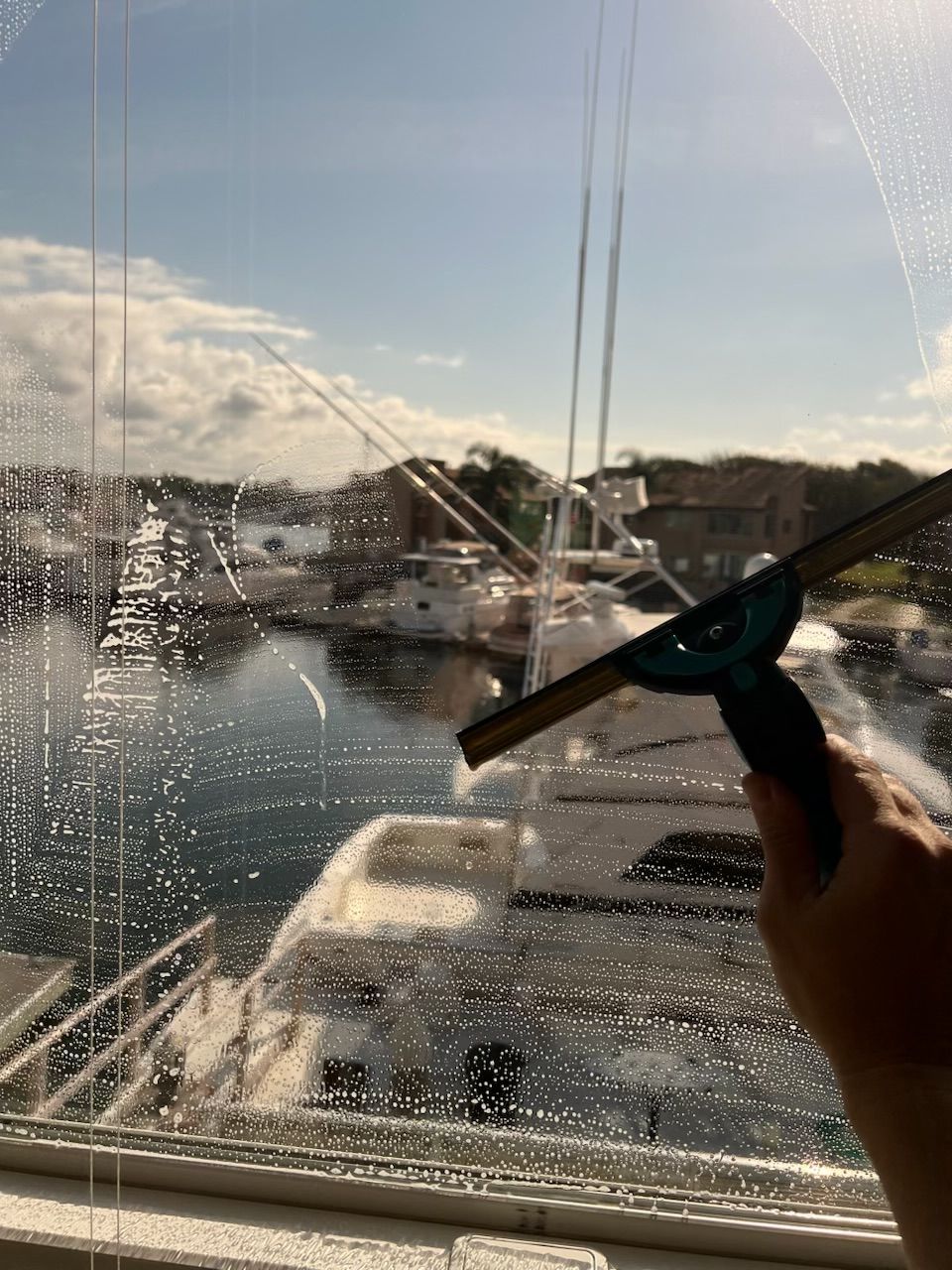 Person cleaning a window with a squeegee, overlooking a marina at sunset.