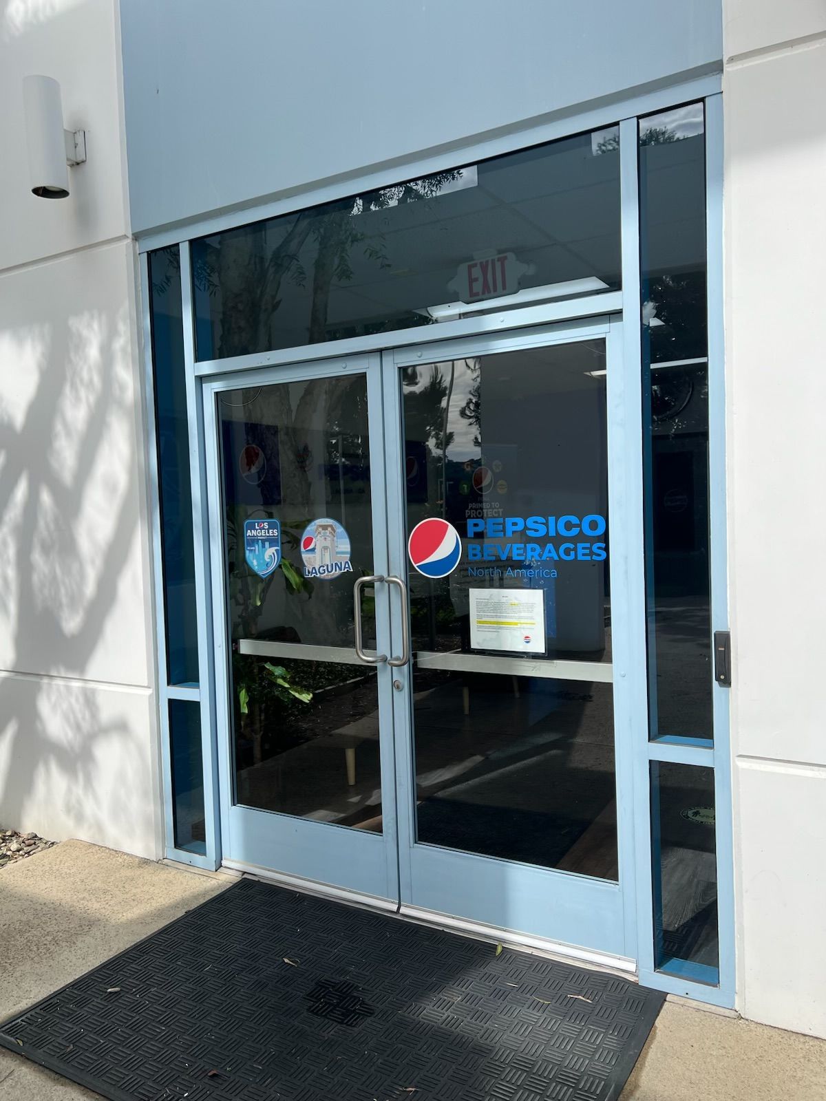 Glass entrance to a building with Pepsi and other decals, framed in blue, beside a sidewalk mat.