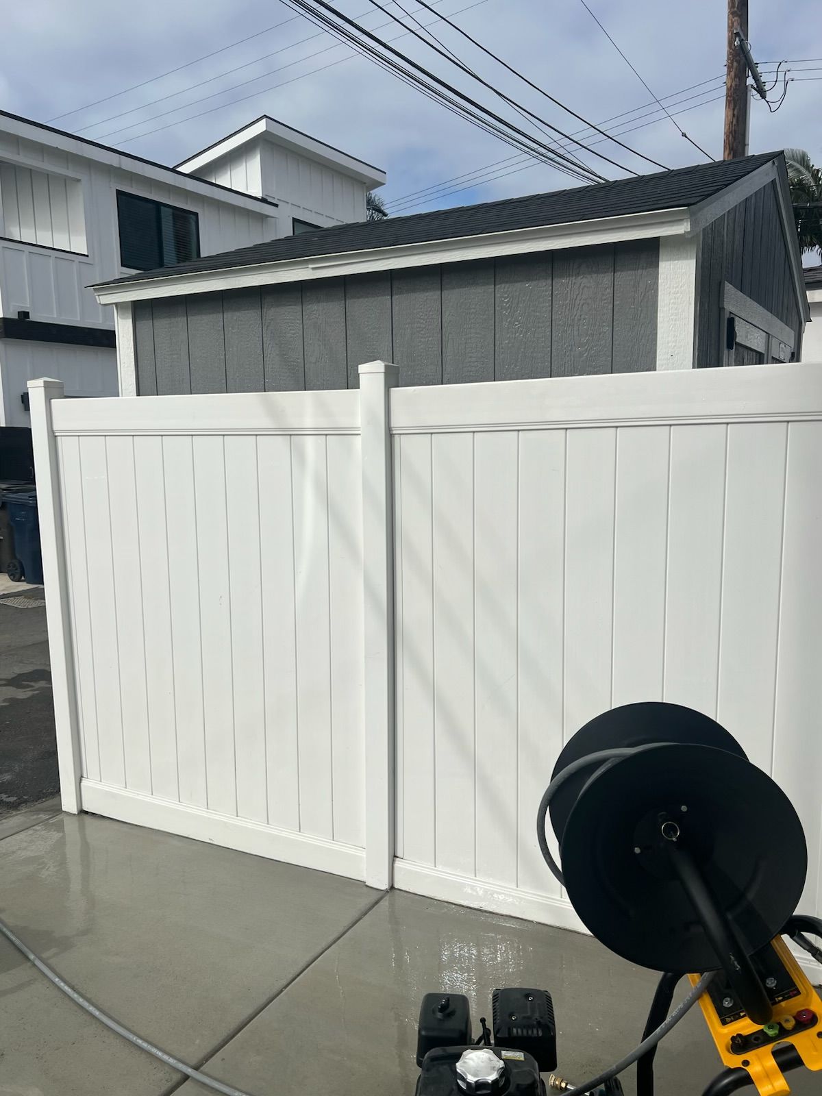 White backyard fence and shed beside a concrete driveway, with a black fire pit in the foreground.