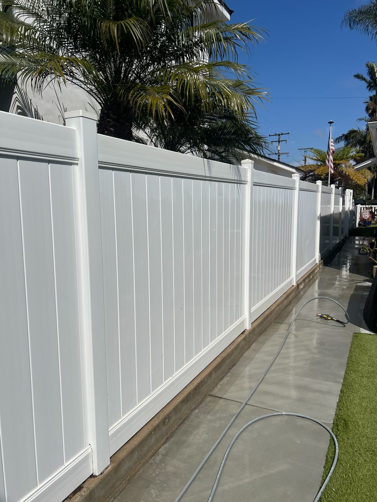 White fence along a sunny sidewalk beside a grassy yard, with palm trees overhead and a hose on the pavement.