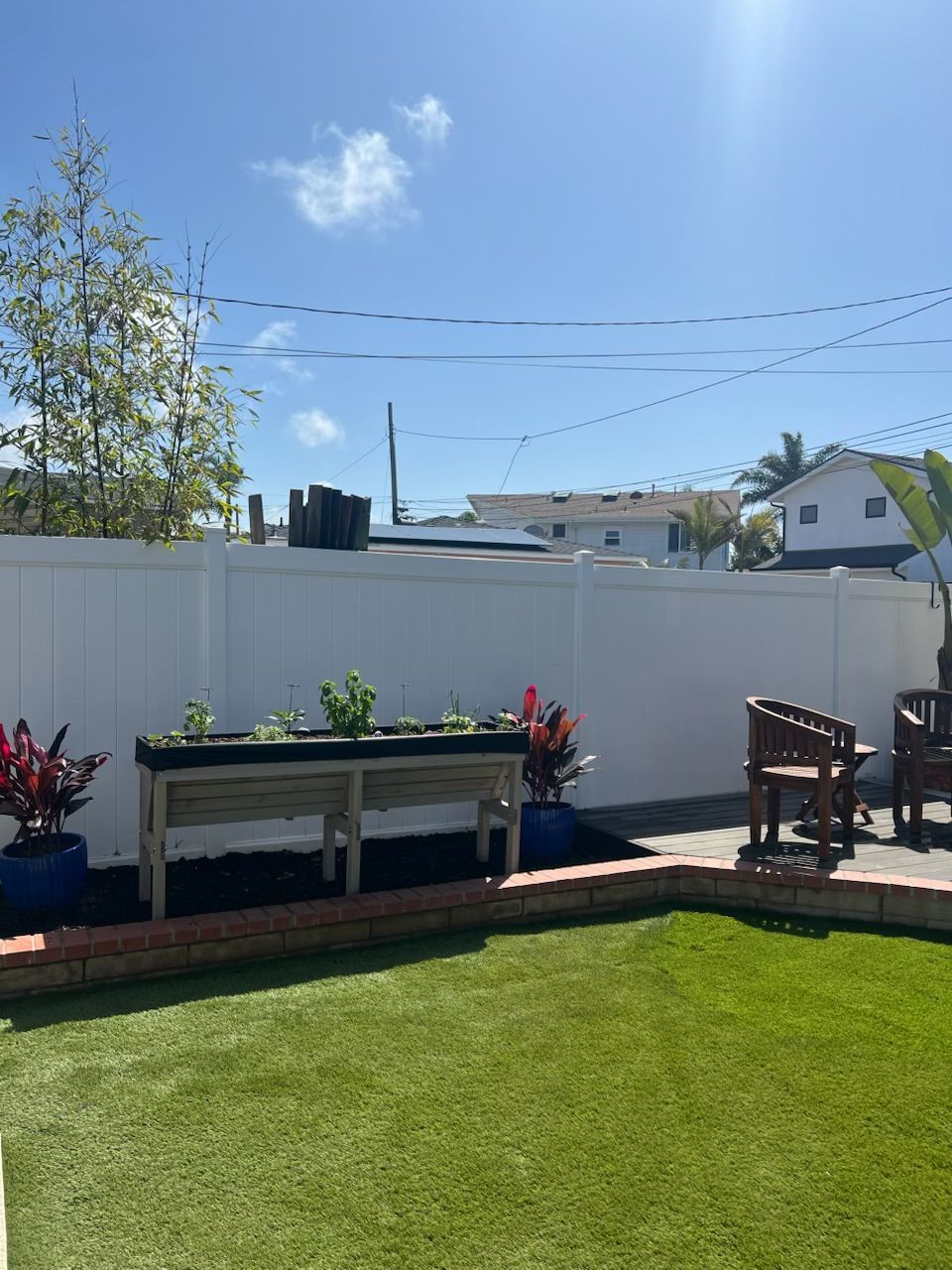 Sunny backyard with green lawn, white fence, raised planter, and two wooden chairs
