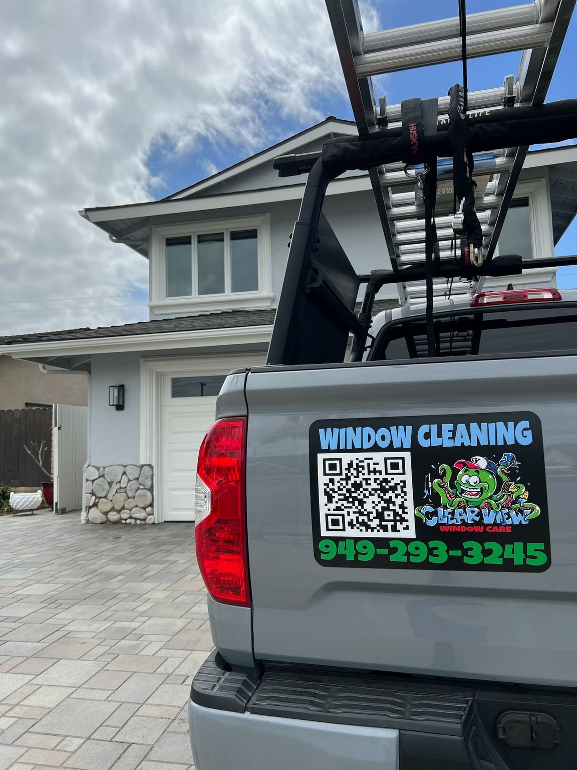 Window cleaning truck parked by a house, with a ladder and bright company decal on the tailgate.