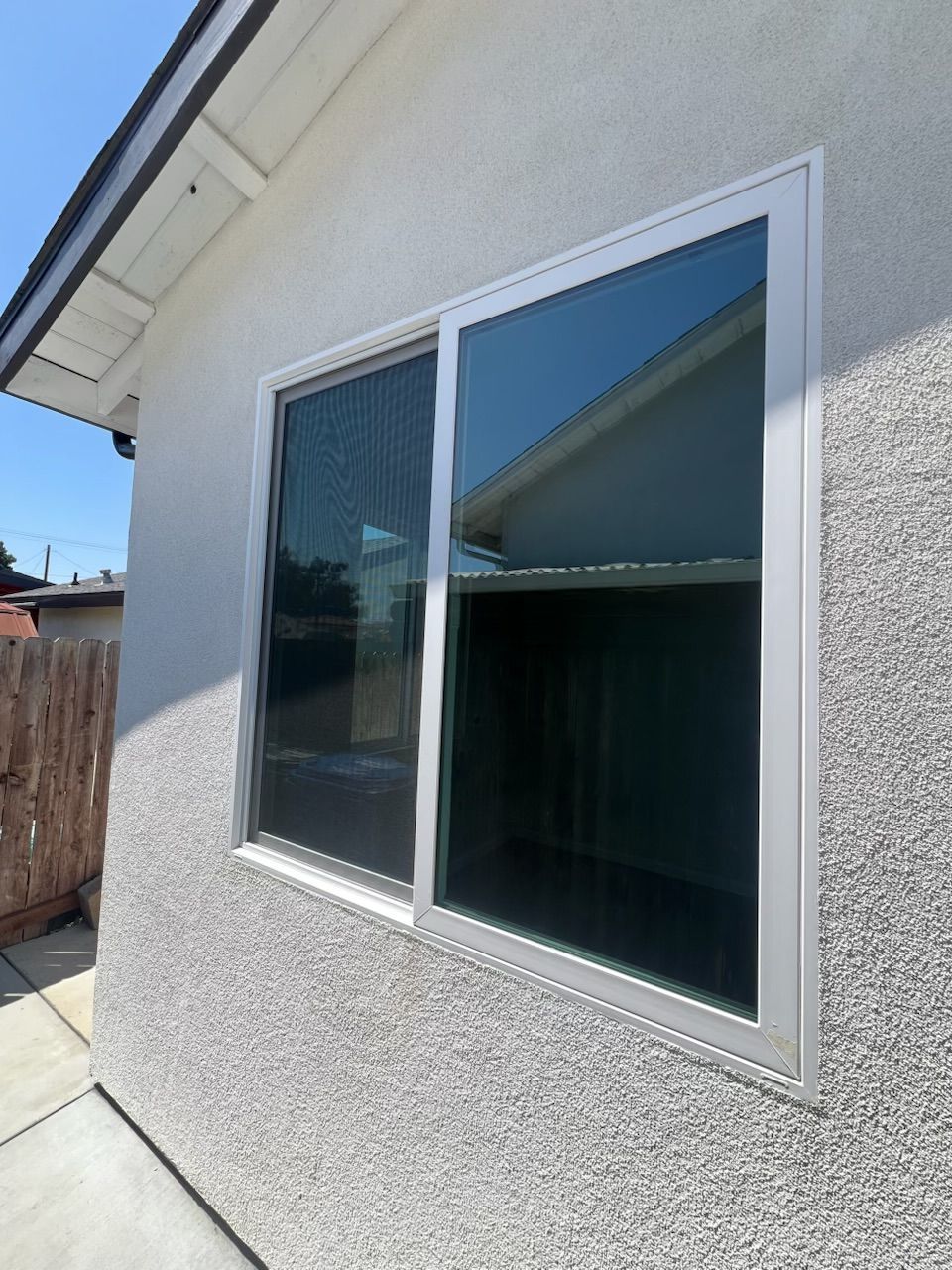 Exterior stucco wall with a white-trimmed sliding window reflecting a roof and blue sky