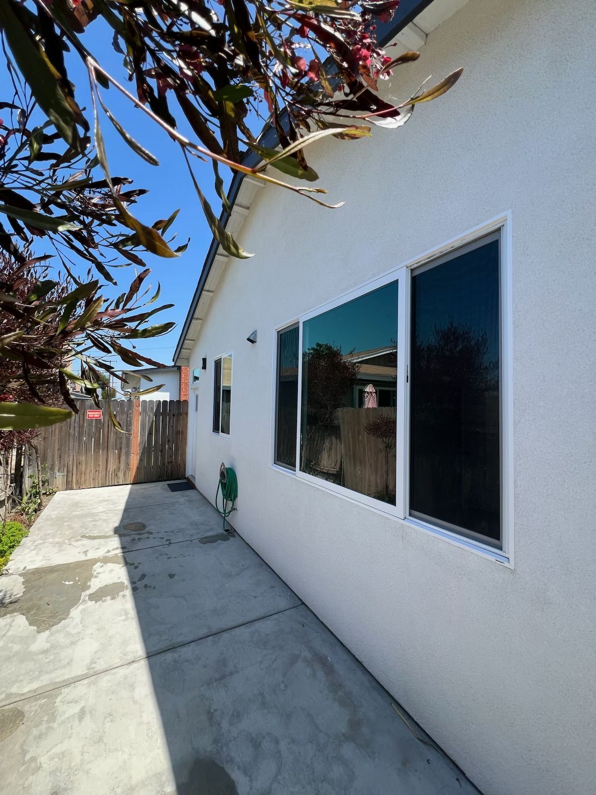 White stucco house exterior with large dark windows and a concrete side walkway under tree branches.