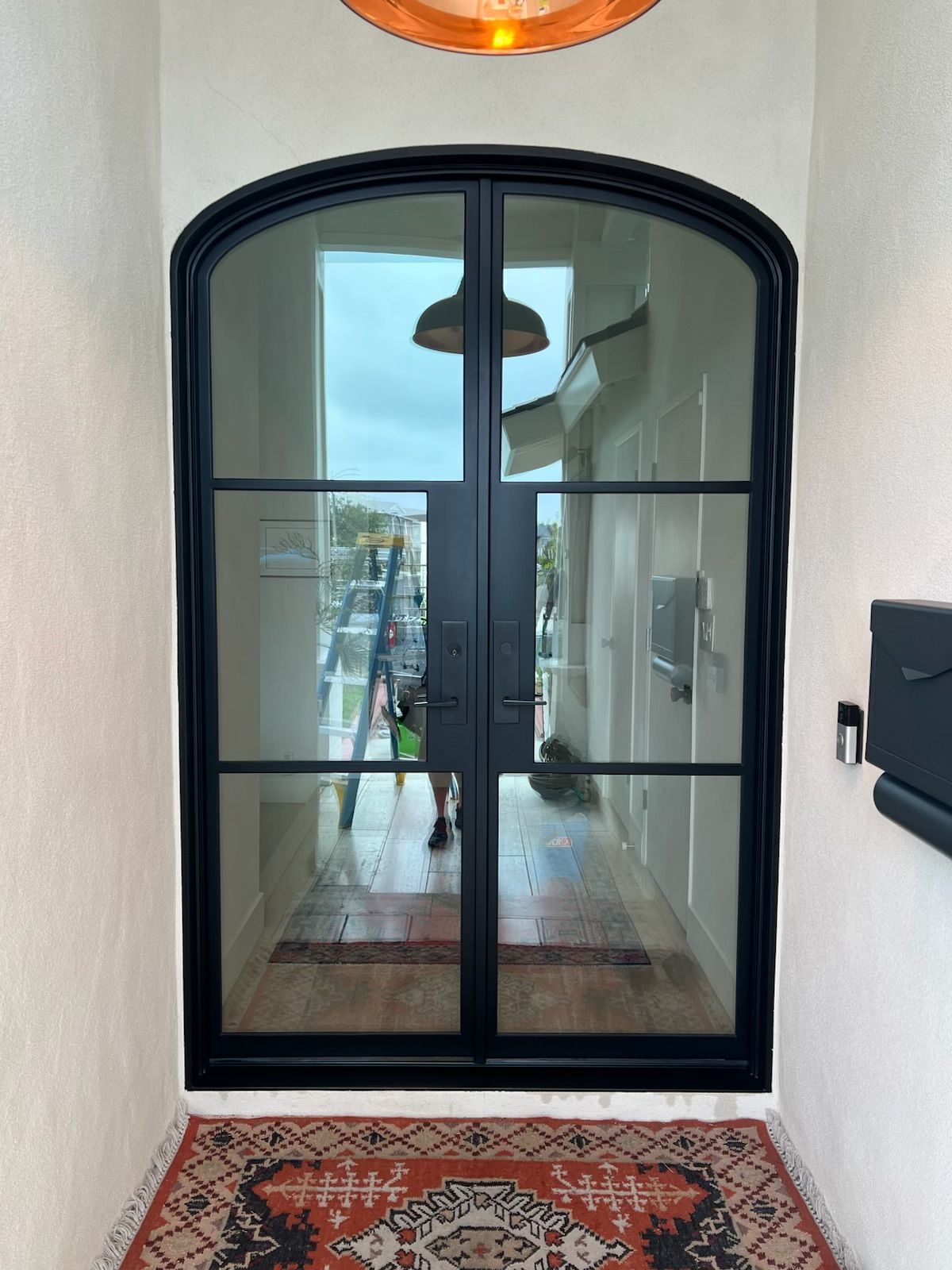 Black arched double glass doors in a white hallway with a patterned rug and ceiling light reflection