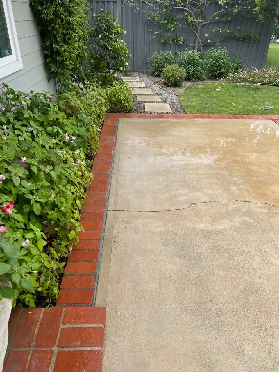 Concrete patio with red brick border beside lush garden and stepping-stone path to a fence.