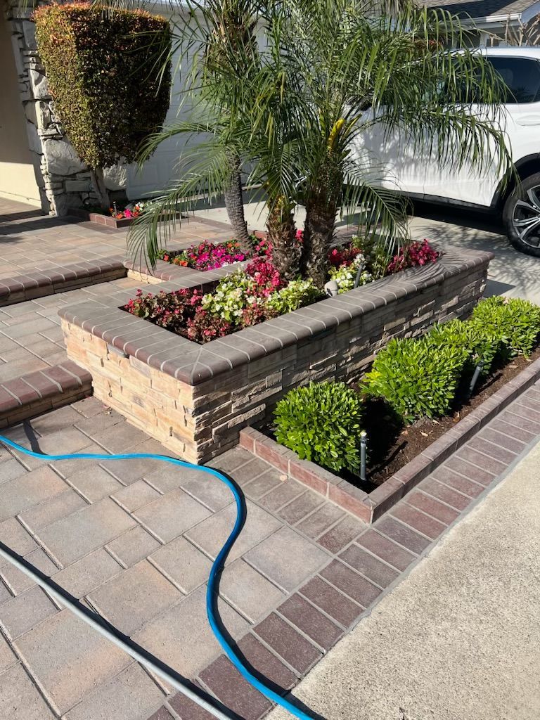 Front yard brick planter with flowers, palms, and trimmed shrubs beside a driveway and parked white car