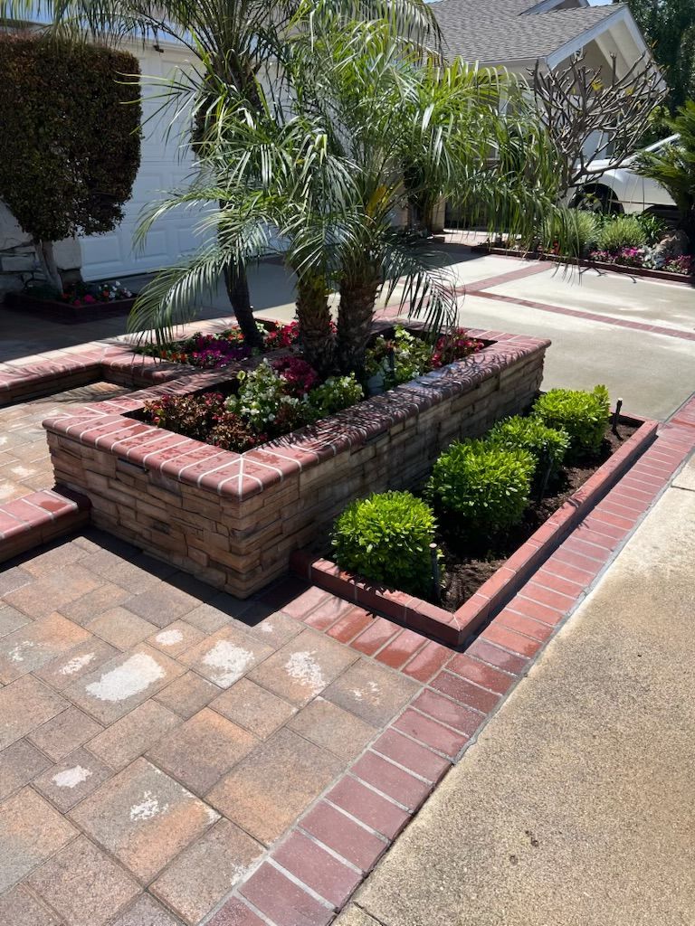 Brick planter with palm trees and red flowers beside a paved driveway in front of a house