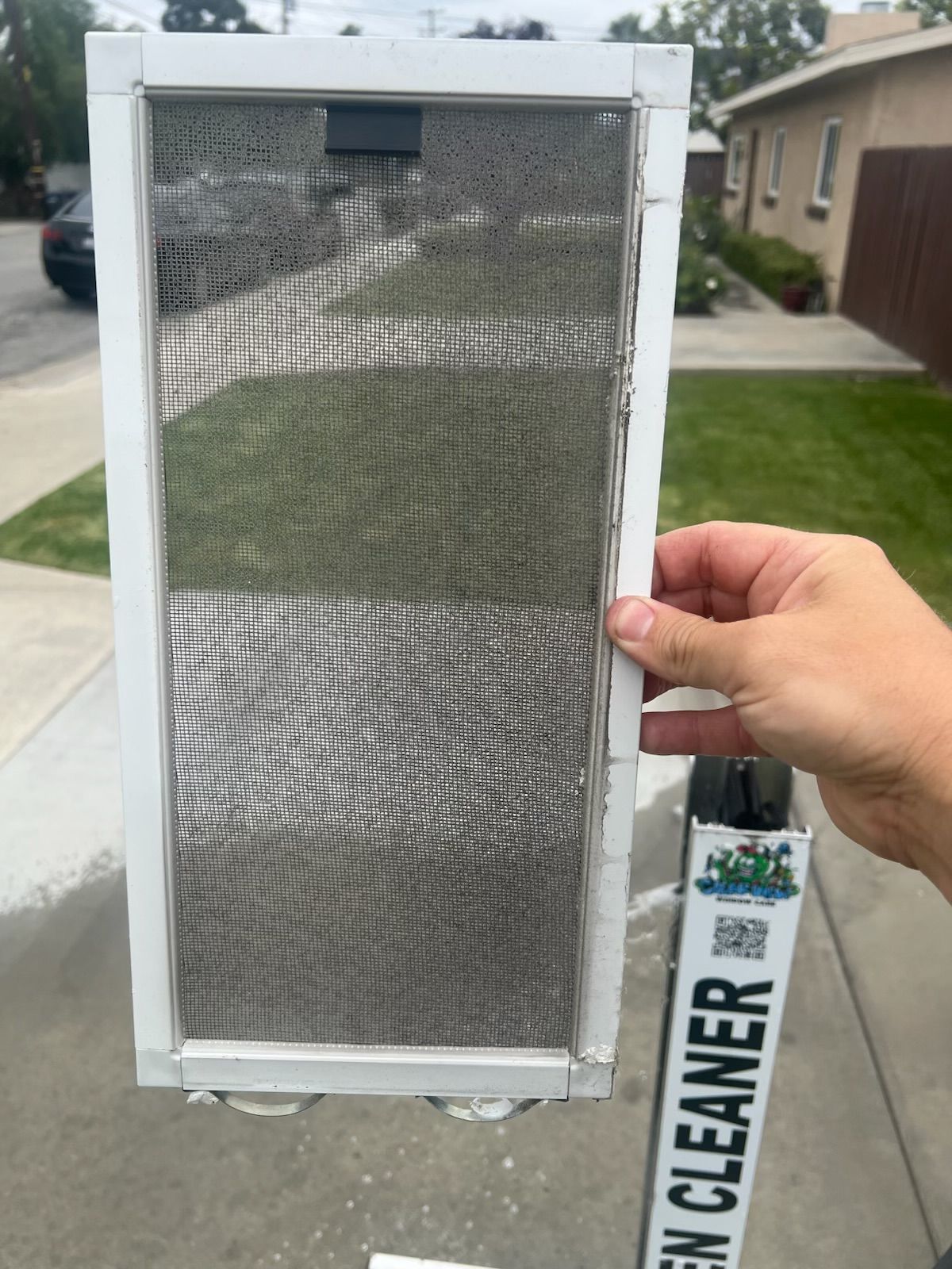 Handheld white metal air filter or vent screen with mesh, outdoors near a house and driveway