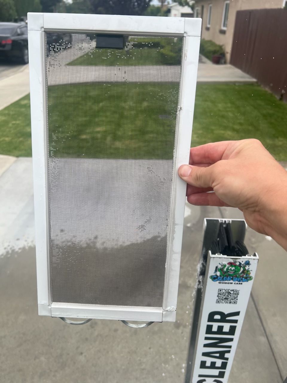 Handheld window screen with white frame and torn mesh beside a window screen cleaner box outdoors