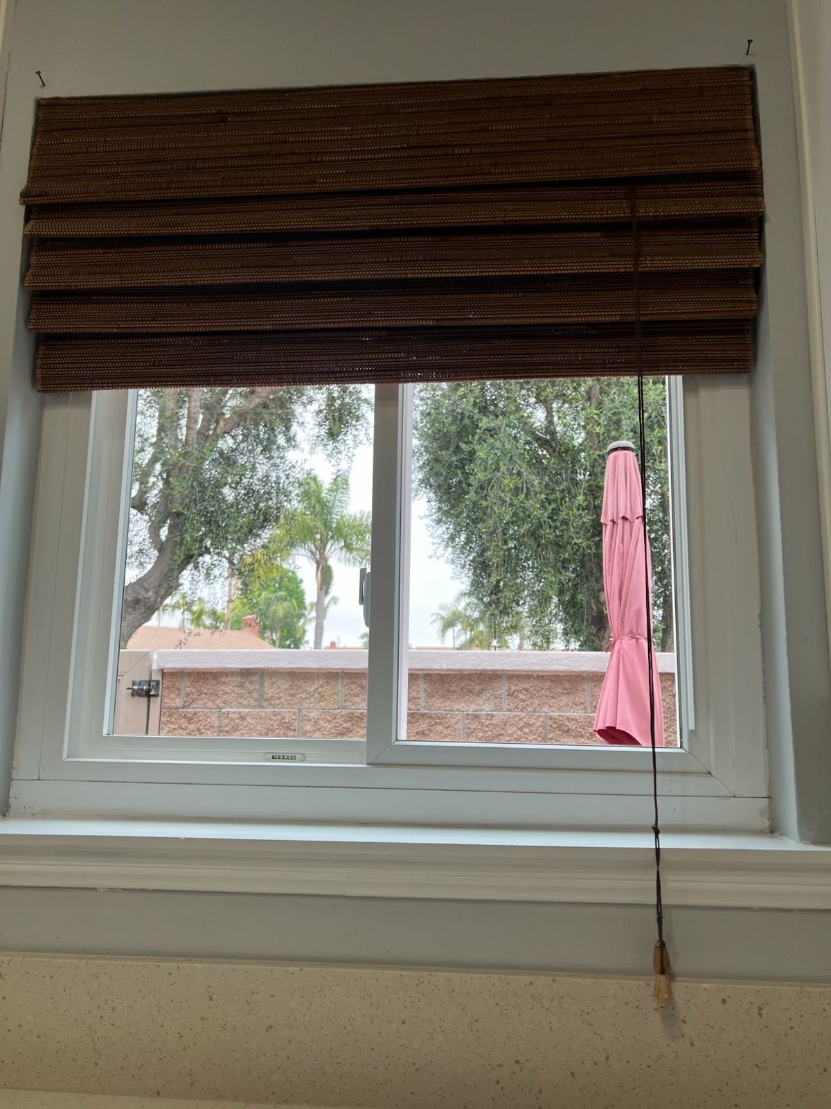 Window with brown blinds, a view of trees and a pink patio umbrella outside