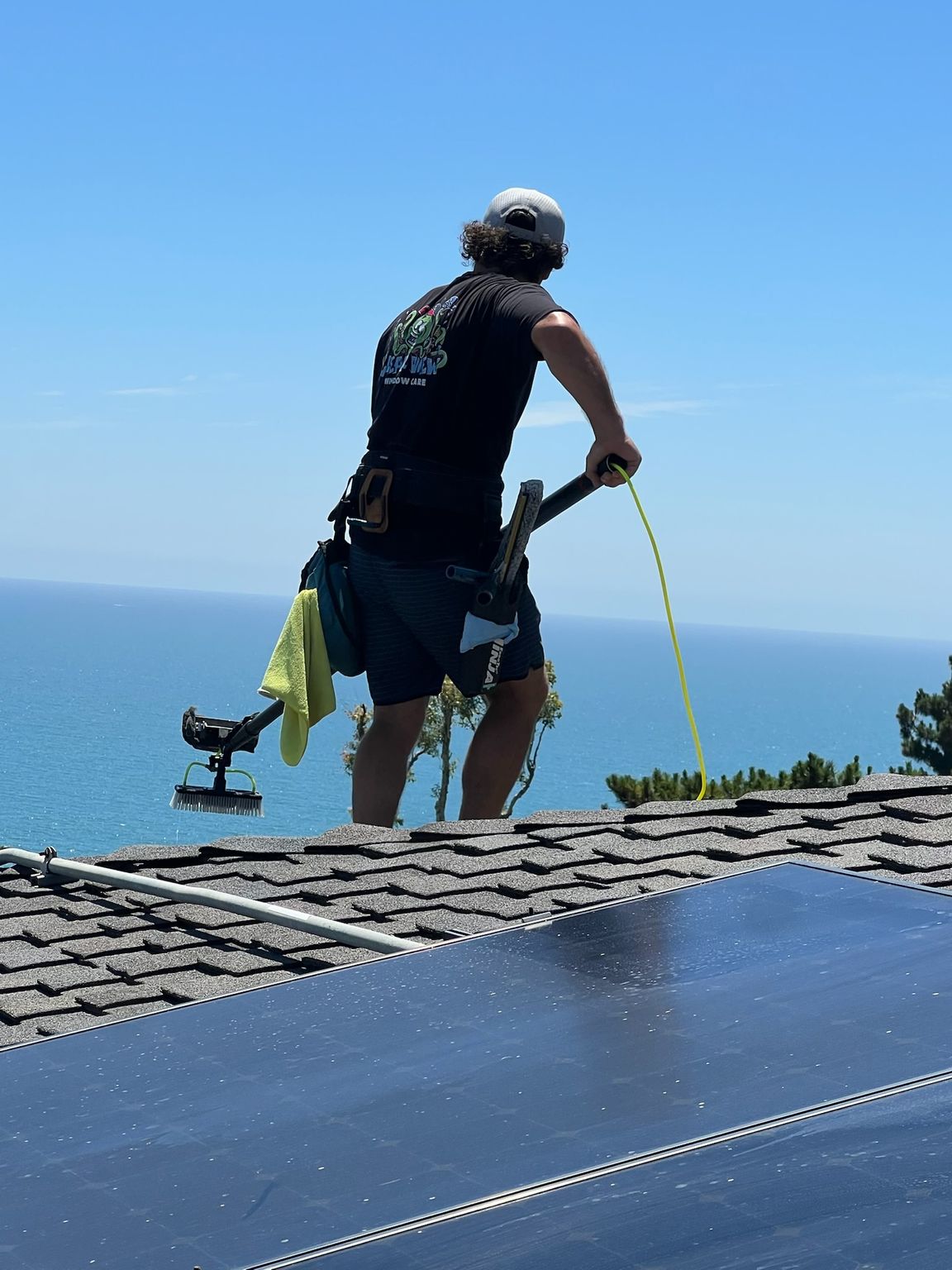 Person on a rooftop using a hose beside a solar panel, with ocean horizon in the background.