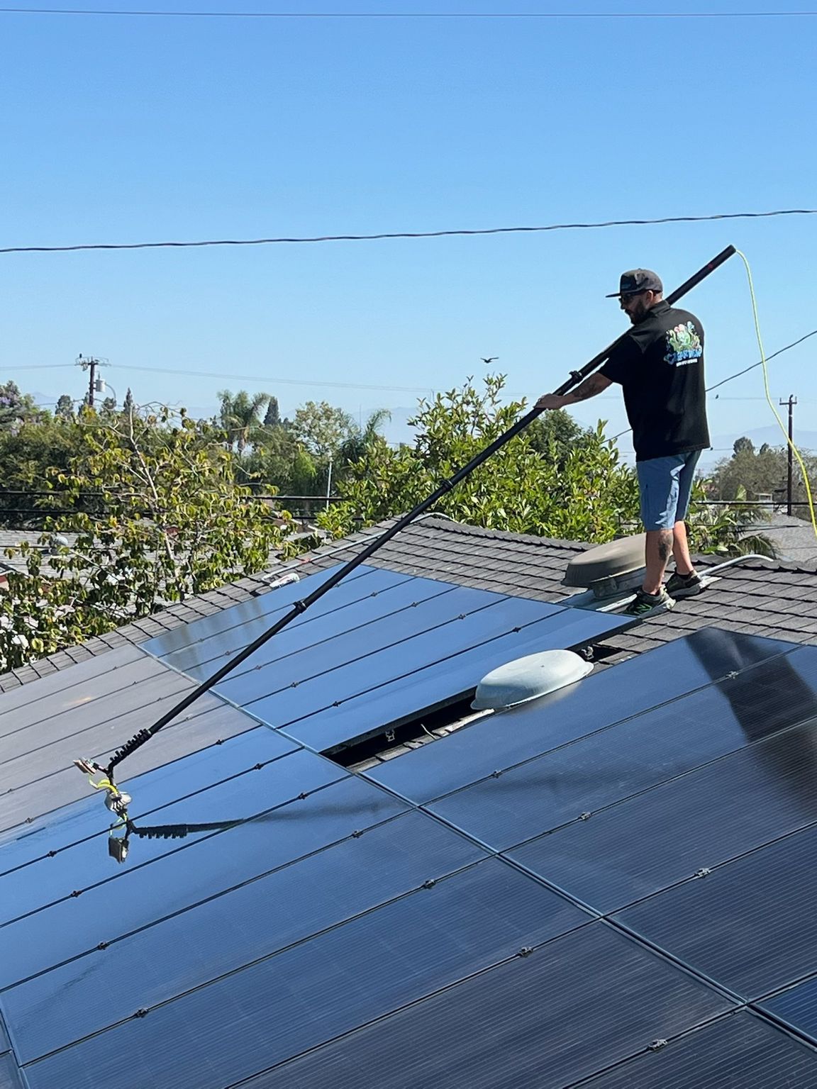 Worker on a solar panel roof using a long pole under a clear blue sky.