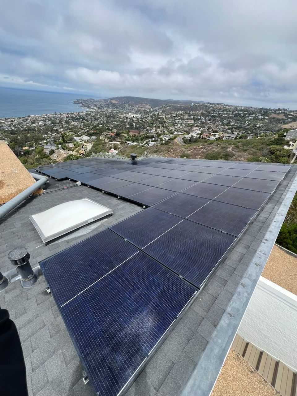 Solar panels on a rooftop overlooking a coastal hillside neighborhood under cloudy skies.