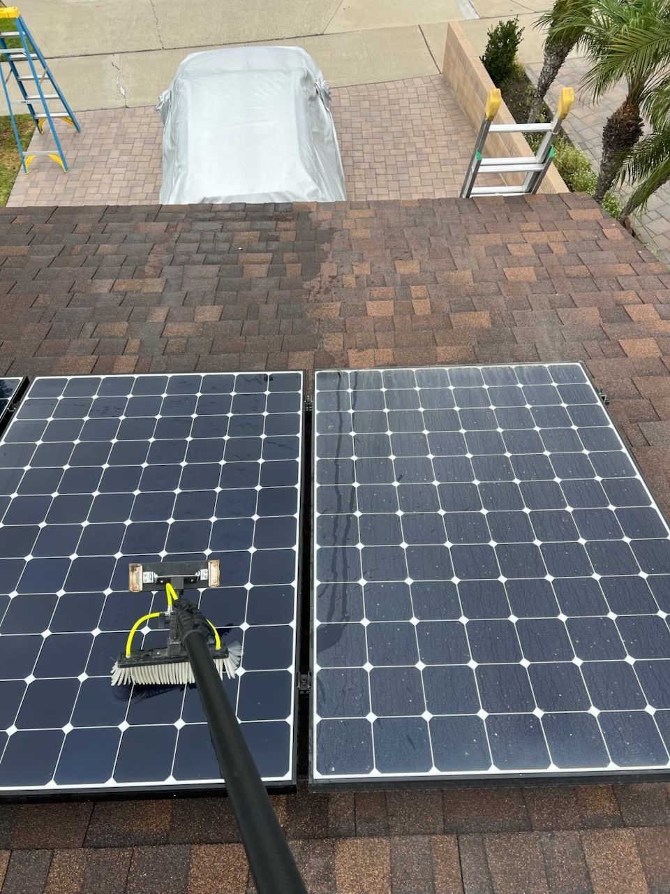 Roof-mounted solar panels being cleaned with a long-handled tool on a shingled roof.