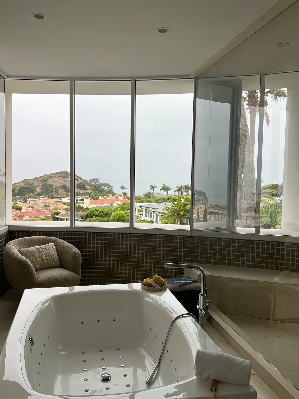 Spa bathroom with a soaking tub, wicker chair, and large windows overlooking the ocean and hills