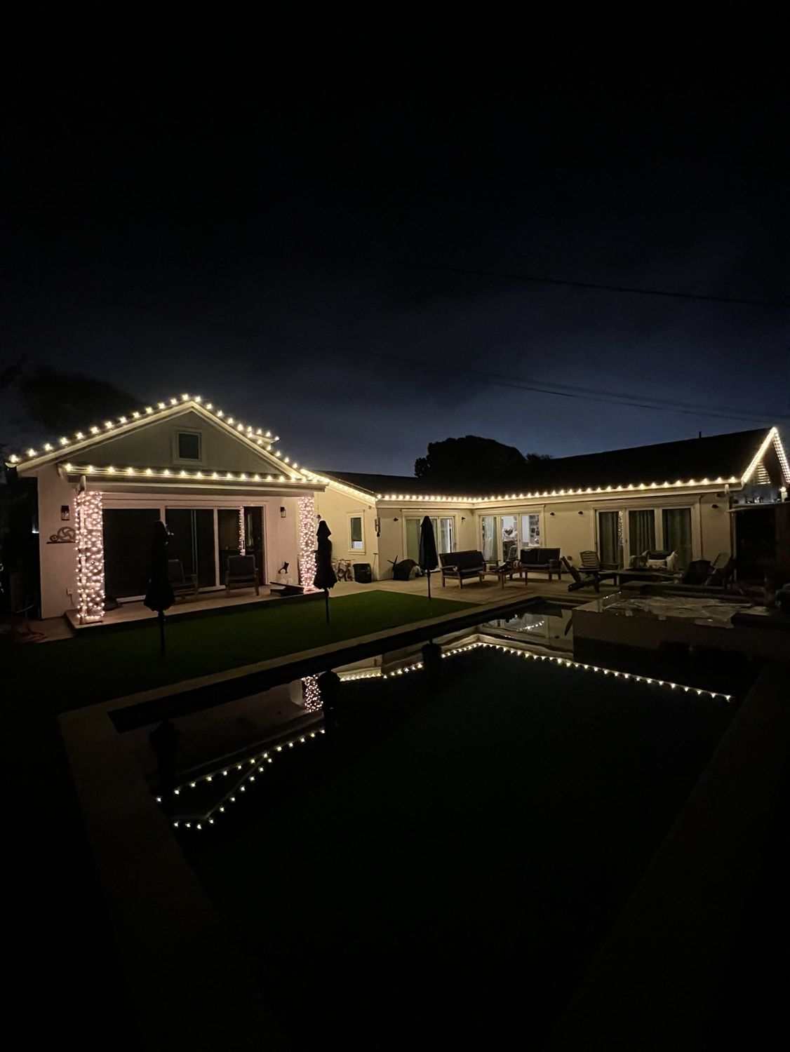 Lit backyard patio at night with string lights reflecting in a pool under a dark sky