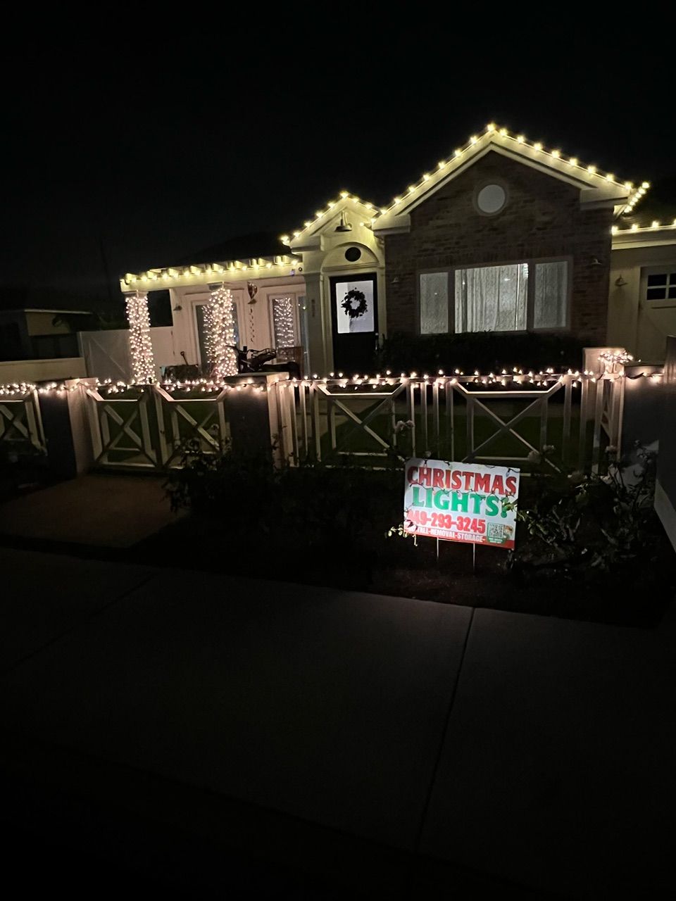House at night outlined with white Christmas lights and a colorful “Christmas Lights” yard sign in front