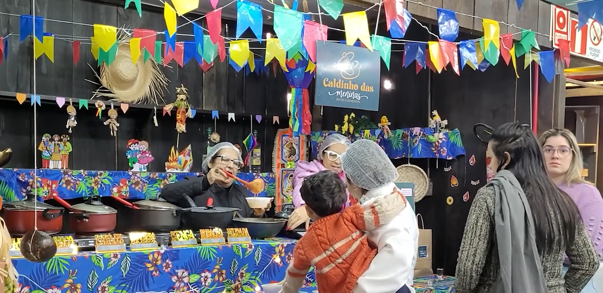 Barraca de jogos de carnaval colorida com balões, bichos de pelúcia e pessoas passeando sob bandeirinhas brilhantes.