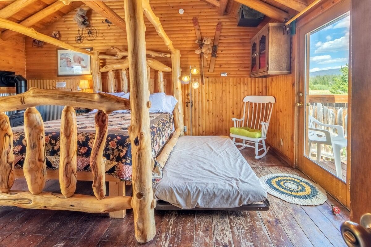 Rustic log cabin bedroom with wood beams, a bed, rocking chair, and large window overlooking mountains