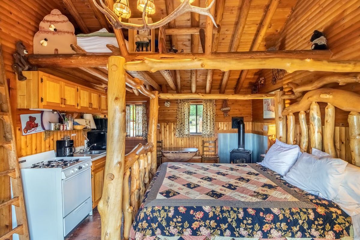 Rustic log cabin bedroom with wooden beams, bed, stove, and small kitchen area