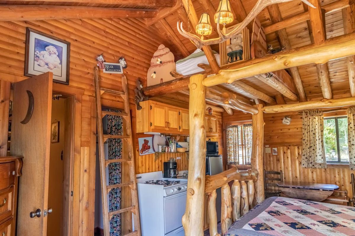 Rustic log cabin interior with wooden beams, small kitchen nook, and a bed by the window