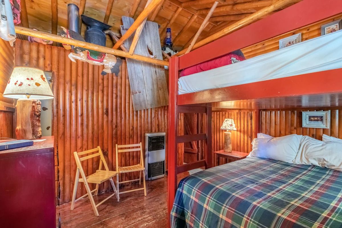 Cozy rustic bedroom with wood-paneled walls, plaid bedspread, red post bed, and two folding chairs