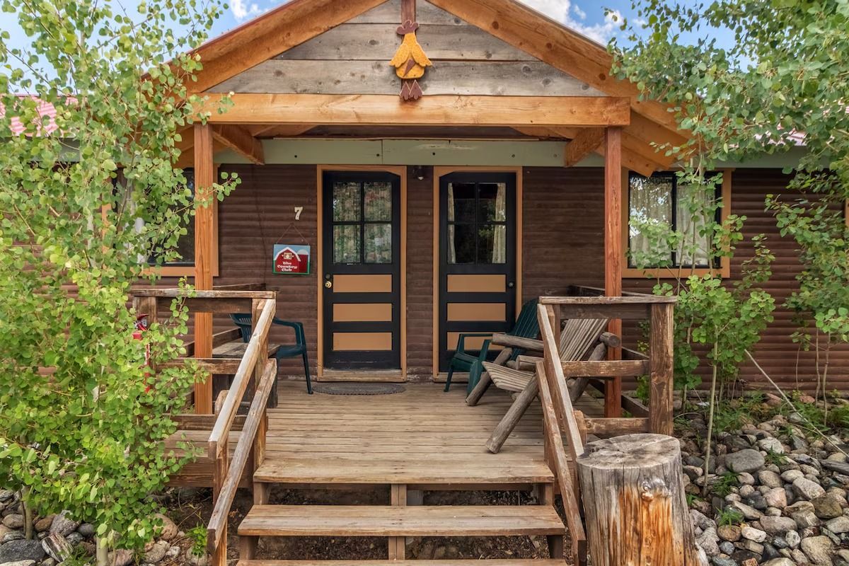 Rustic cabin porch with wooden steps, railings, and double glass doors surrounded by trees