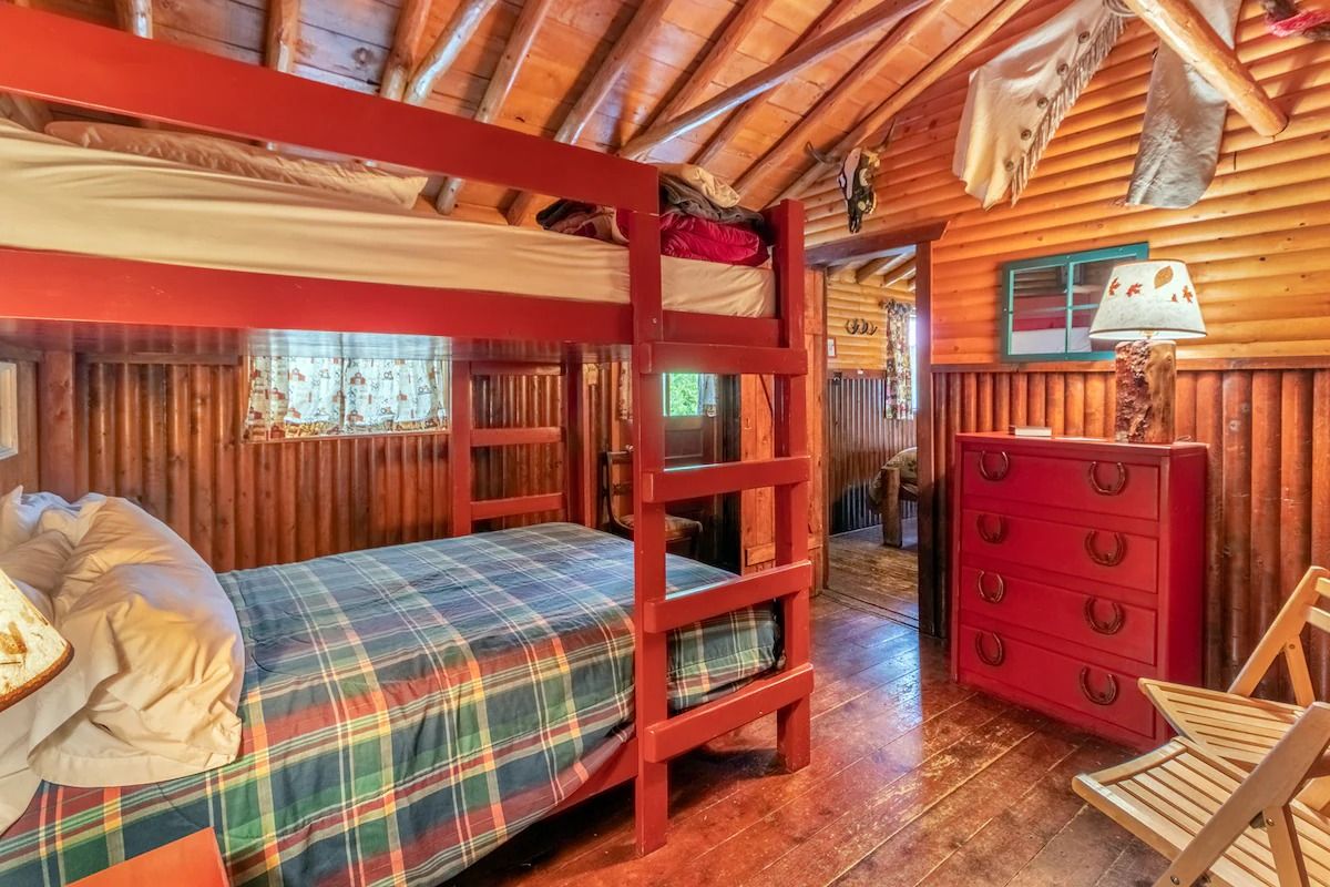 Rustic log cabin bedroom with red bunk bed, plaid bedding, and red dresser under warm wood beams
