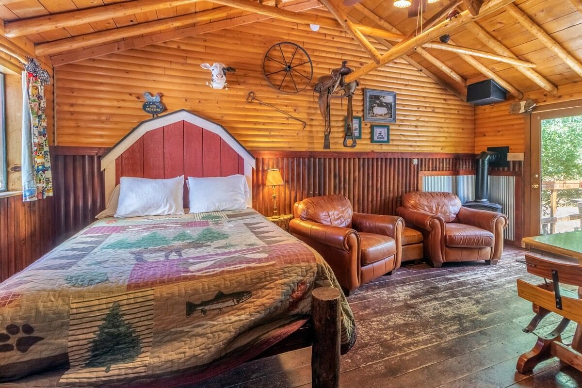 Rustic cabin bedroom with wood-paneled walls, log ceiling, bed, leather chairs, and mountain quilt artwork.