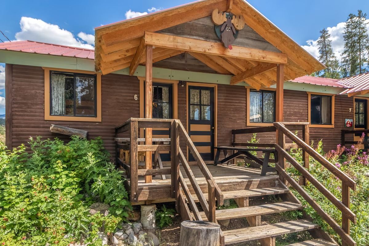 Rustic wooden cabin with front porch, steps, and green shrubs under a bright sky