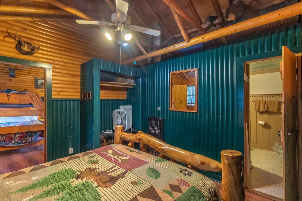 Rustic log cabin bedroom with teal walls, wooden ceiling, quilted bed, and bathroom doorway visible