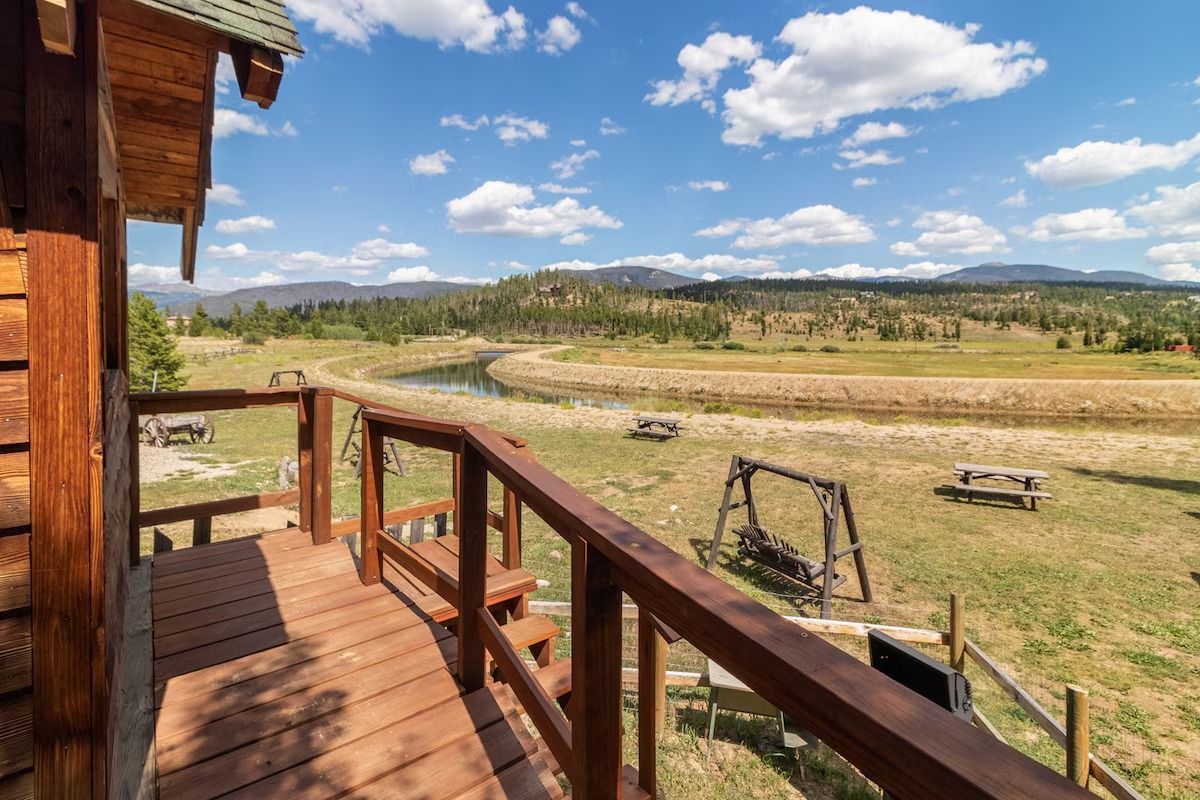 Wooden cabin porch overlooking a sunny grassy field and distant hills under a blue sky with clouds