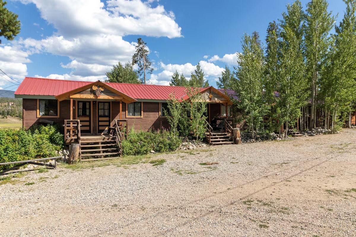 Rustic wooden cabin with red roof, front steps, and trees on a gravel clearing under a blue sky