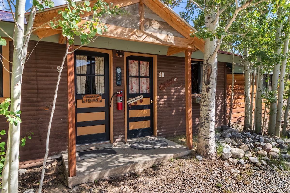 Rustic cabin entrance with double doors, wood siding, and a small porch surrounded by trees and rocks