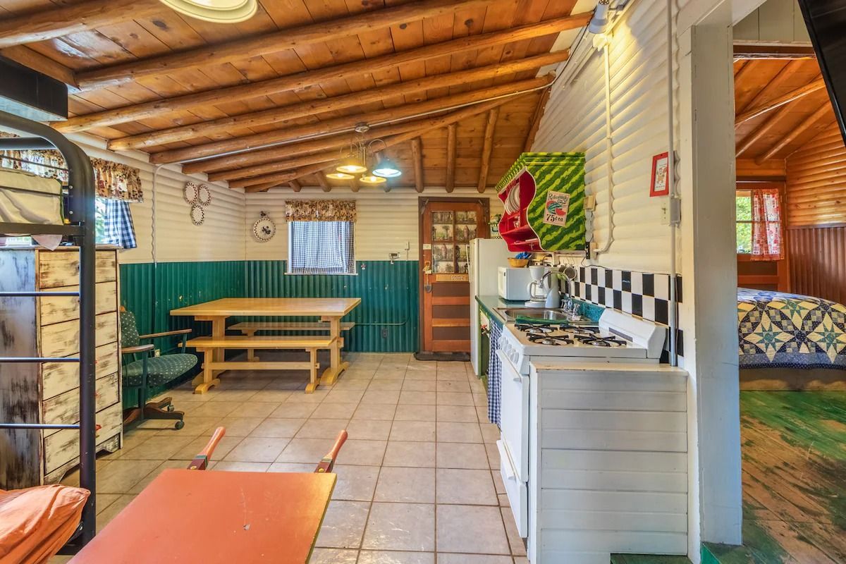 Rustic indoor dining area with wood-beam ceiling, picnic table, tiled floor, and green curtains.