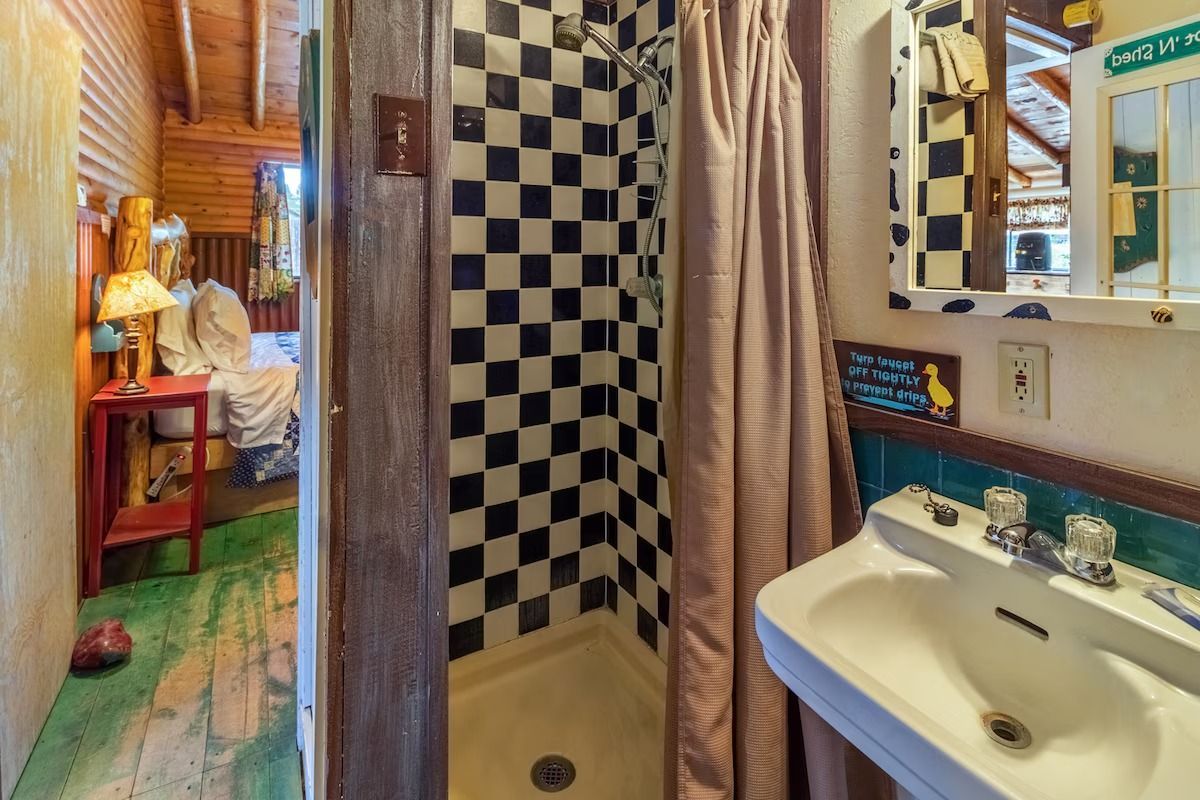 Narrow bathroom with checkerboard shower, sink, and open doorway to a rustic room with a bed.