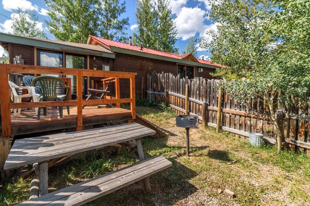 Wooden porch and deck with stairs, fenced yard, grill, and trees under a partly cloudy sky