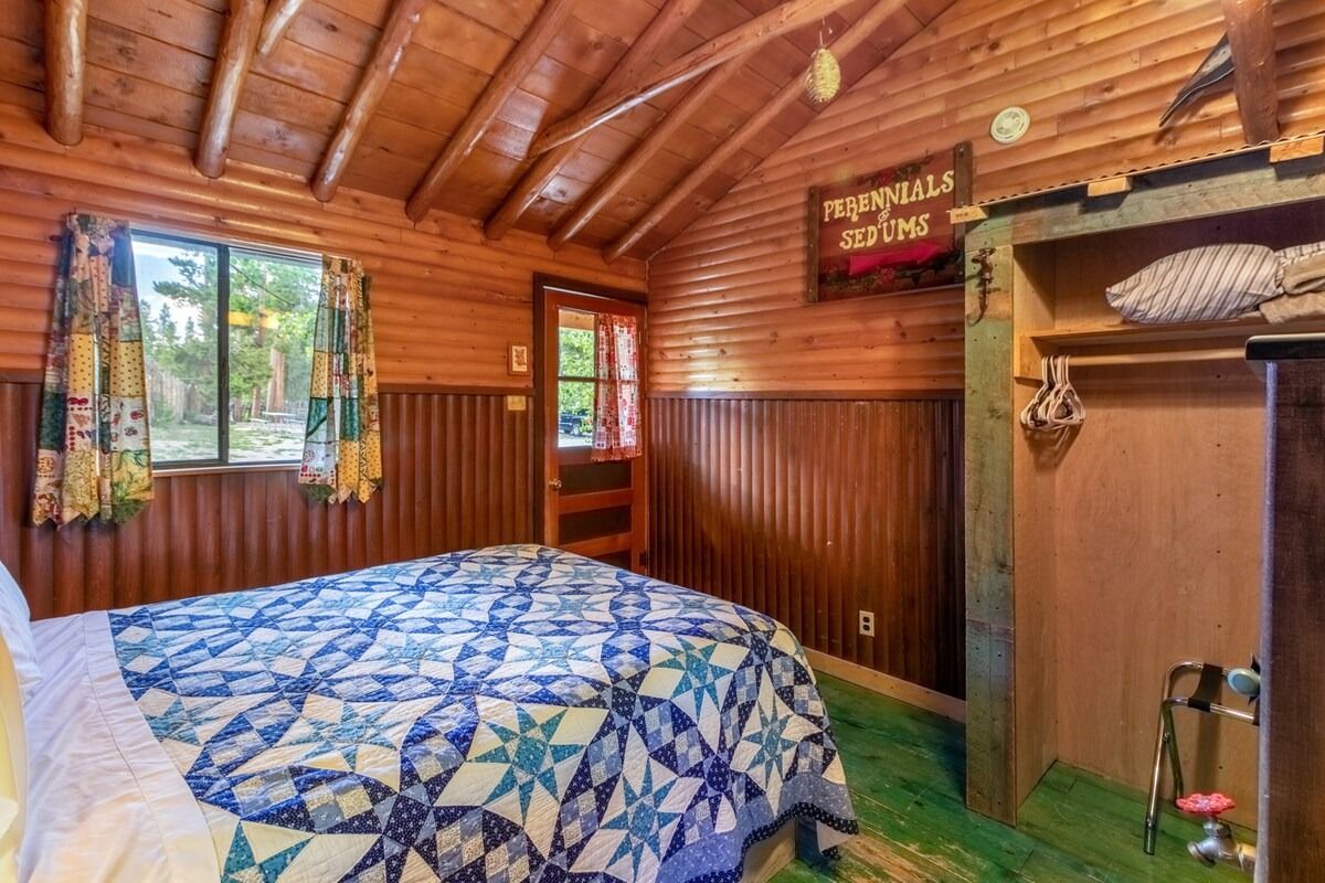 Wood-paneled cabin bedroom with blue patterned bedspread and two windows with colorful curtains