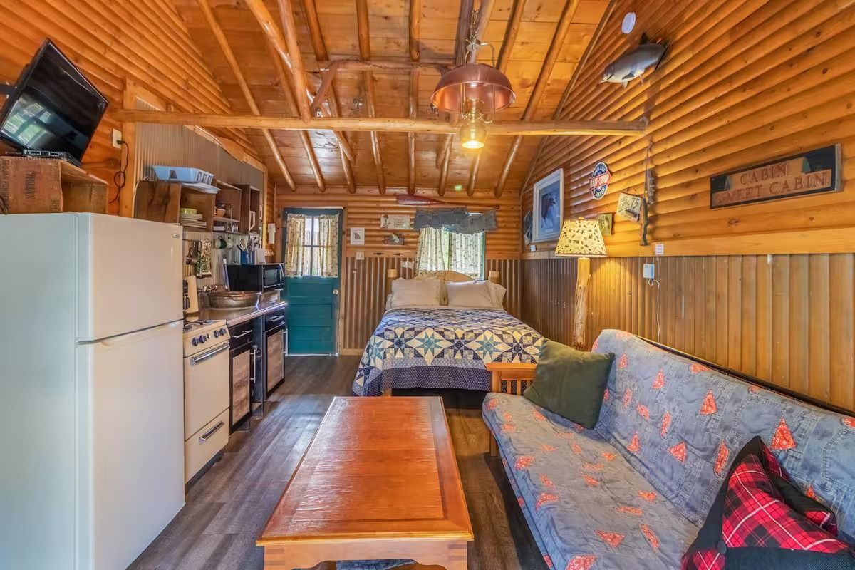 Cozy rustic cabin kitchen and living area with wood-paneled walls, sofa, table, and white refrigerator
