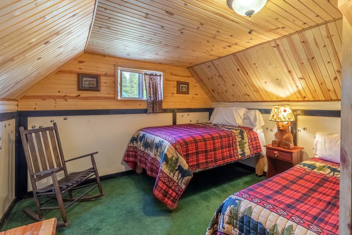 Cozy attic bedroom with two plaid-covered beds, green carpet, wood-paneled sloped ceiling, and lamp lit table