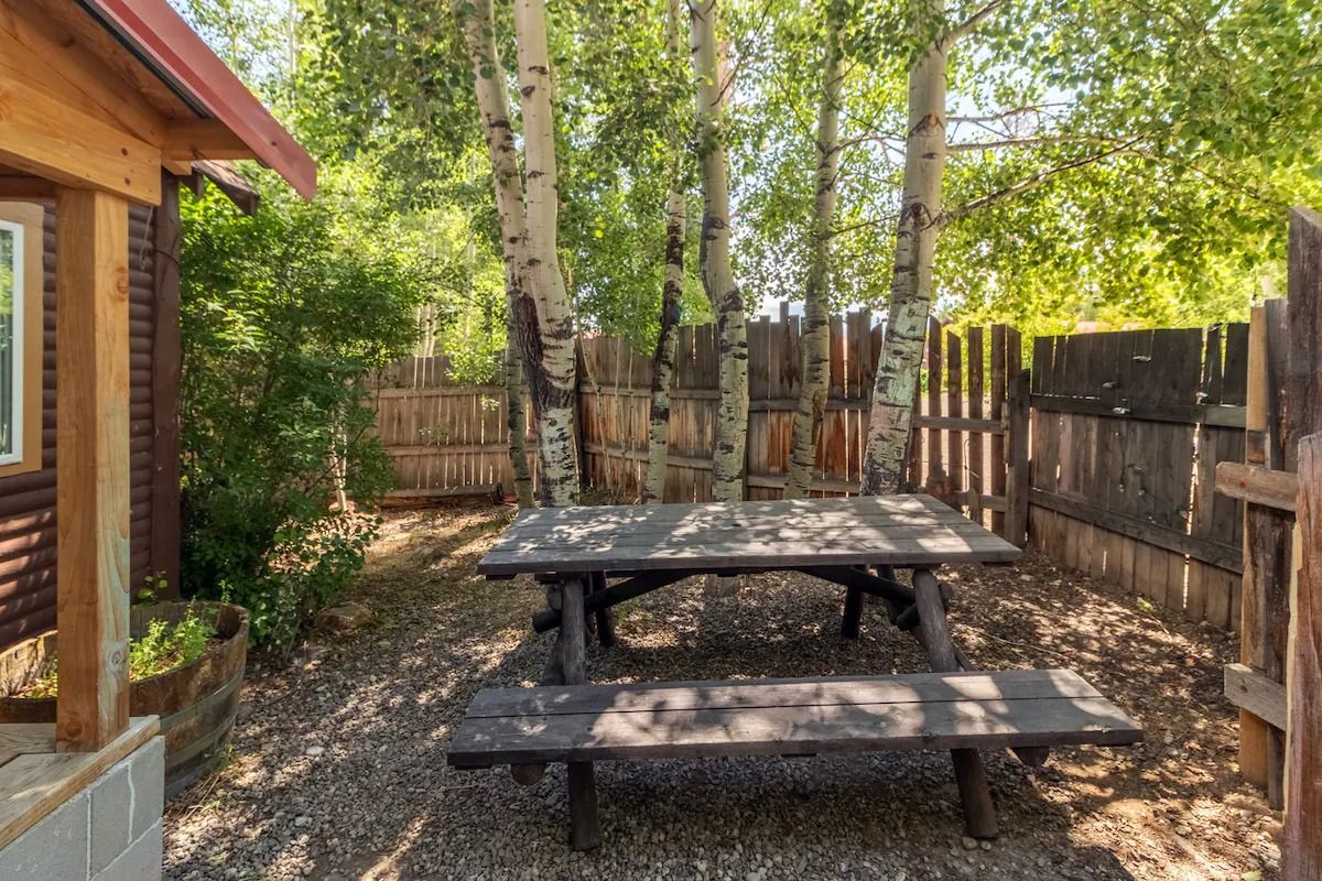 Wooden picnic table in a shaded backyard beside a house and fence, surrounded by trees and gravel.